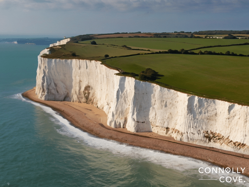 Aerial view of the White Cliffs of Dover, one of England’s natural wonders, with green fields above and a pebble beach below, bordered by the calm blue sea.