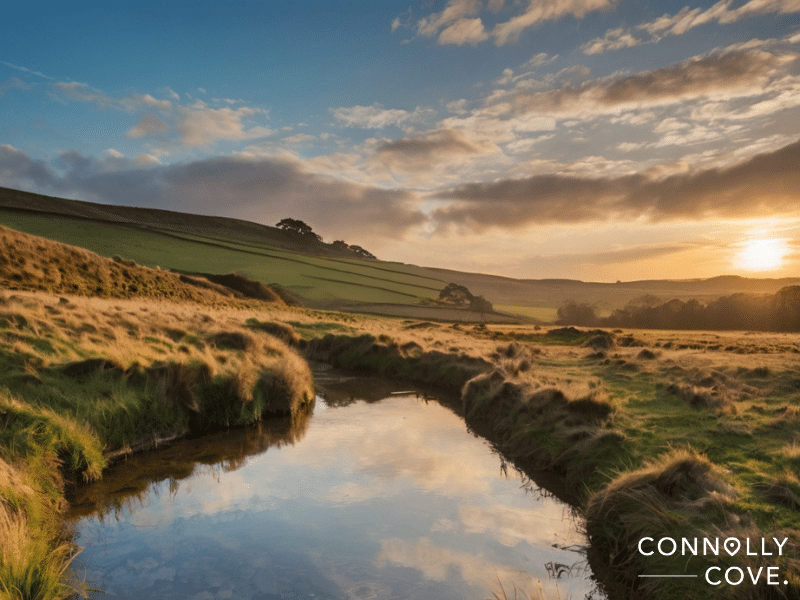 A tranquil countryside landscape at sunset, featuring a reflective stream, grassy fields, rolling hills, and a partly cloudy sky with the sun low on the horizon—perfect for a peaceful retreat in England. CONNOLLY COVE text appears in the corner.