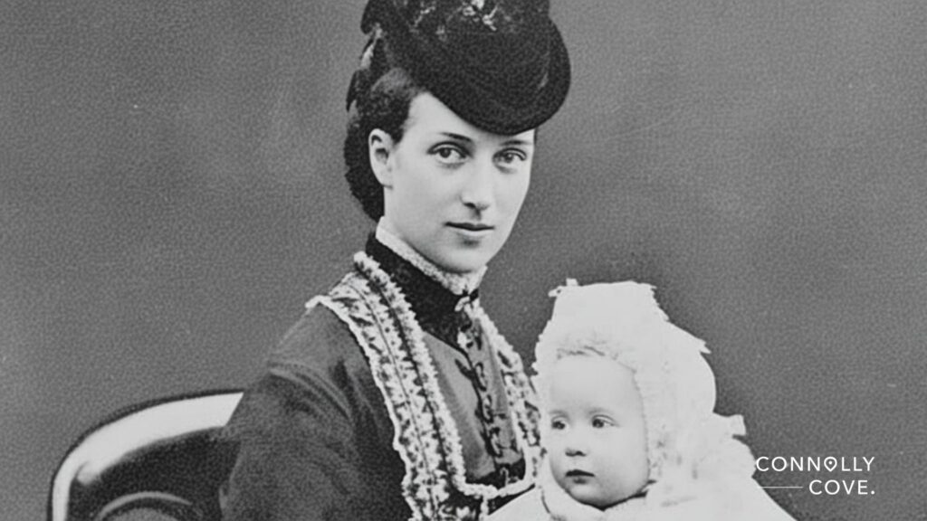 A black-and-white photo of a woman in Victorian clothing and hat sitting on a chair, holding a baby in a bonnet and gown. Echoing the era of Victoria, the plain background highlights their timeless bond. CONNOLLY COVE. appears at bottom right.