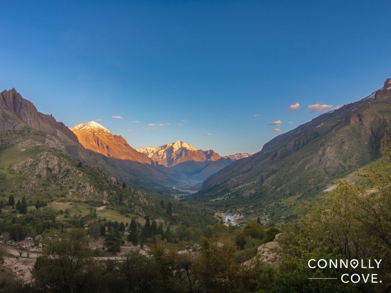 A scenic mountain valley at sunset in Western Asia, with snow-capped peaks glowing orange, green hills, trees, and a winding river below; the sky is mostly clear. Connolly Cove is written in white text in the bottom right corner.