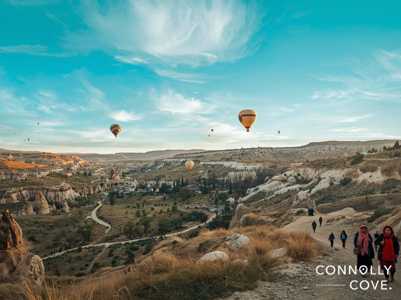 Colorful hot air balloons float over a scenic rocky valley in Western Asia at sunrise, with people walking on a trail in the foreground. The landscape features unique rock formations and sparse vegetation. The logo “Connolly Cove” appears in the corner.