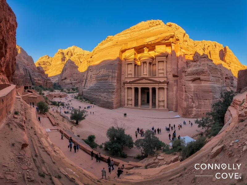 A wide view of Petra’s Al-Khazneh (The Treasury) carved into red rock cliffs in Western Asia, surrounded by visitors, with dramatic sunlight and a clear blue sky. “Connolly Cove” is written at the bottom right.
