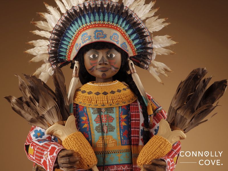 A colorful Native American doll inspired by Hopi kachina dolls, with a feathered headdress, traditional patterned clothing, and painted face holds two ears of corn. The background is brown, and Connolly Cove is written in the corner.