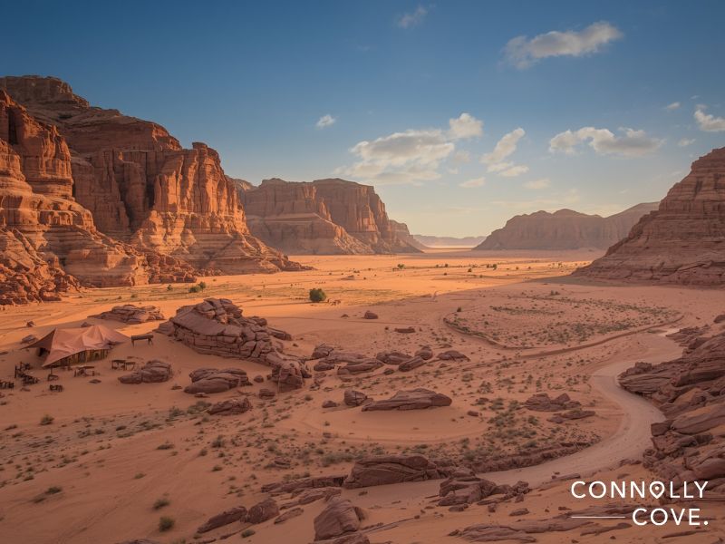 Wide desert landscape with dramatic red rock cliffs, scattered boulders, and sand dunes under a blue sky with clouds evokes the natural beauty of Western Asia. A winding dirt path and a few small structures are visible. Connolly Cove is written in the bottom right corner.