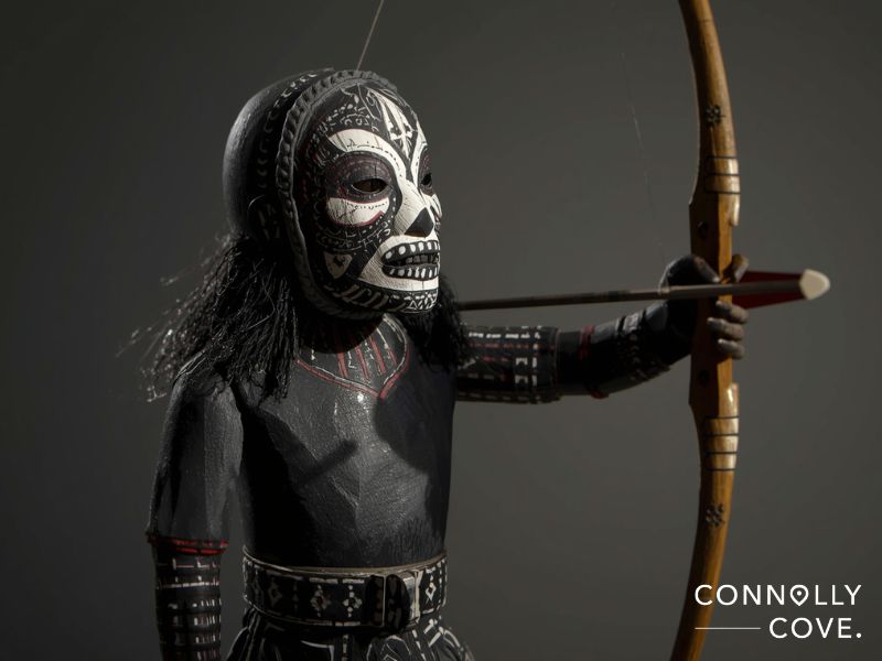 A close-up of a detailed wooden figure inspired by Hopi kachina dolls, with black body paint, a painted tribal mask, and long black hair, drawing a bow with an arrow. The Connolly Cove logo appears in the bottom right corner.