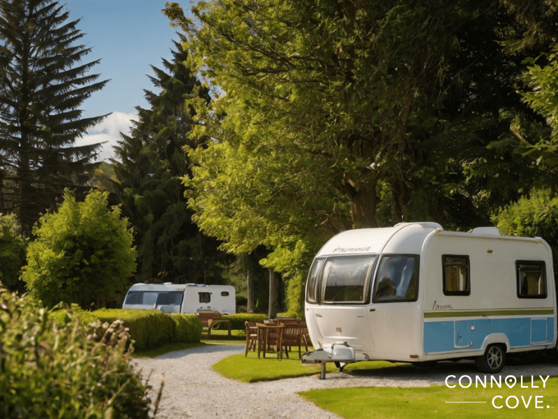 holiday parks in Ireland
Two white caravans are parked on a grassy campsite surrounded by tall trees and hedges. Wooden tables and chairs are set outside, with sunlight filtering through the leaves—a peaceful scene at a Caravan and Camping Park. Connolly Cove is written in the bottom right corner.