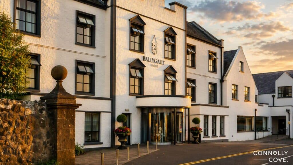 White building with black trim labelled Ballygally Castle Hotel at sunset, featuring arched windows, a round glass entrance, neat landscaping, and stone pillars. The Connolly Cove logo is visible in the corner—a picturesque setting that hints at Ballygally Castle’s history and its famous haunted stories.