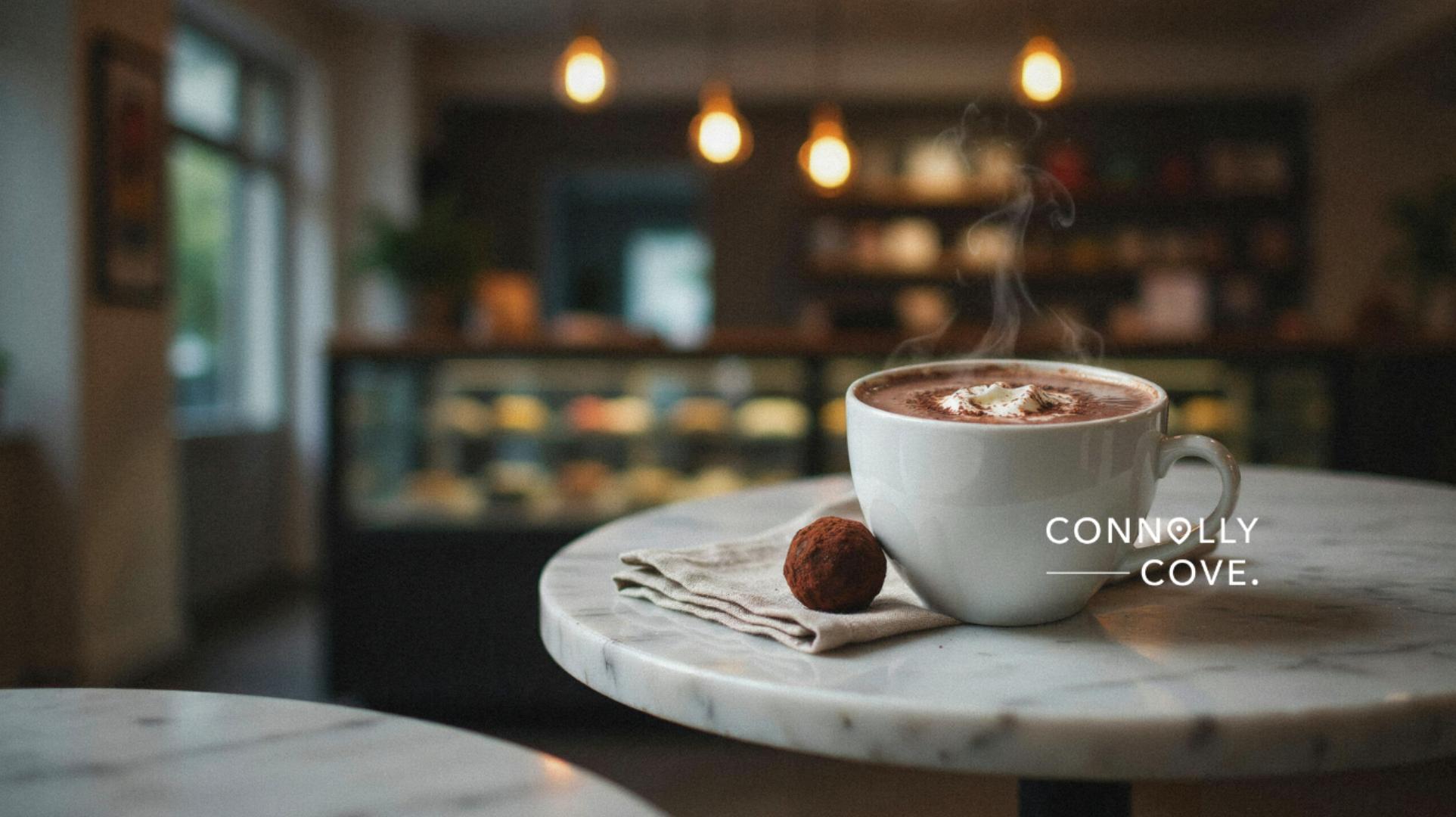 A steaming cup of Co Couture Hot Chocolate with whipped cream and a truffle sits on a marble table in a cosy café, with warm lighting and blurred bakery shelves in the background. The text reads Connolly Cove.