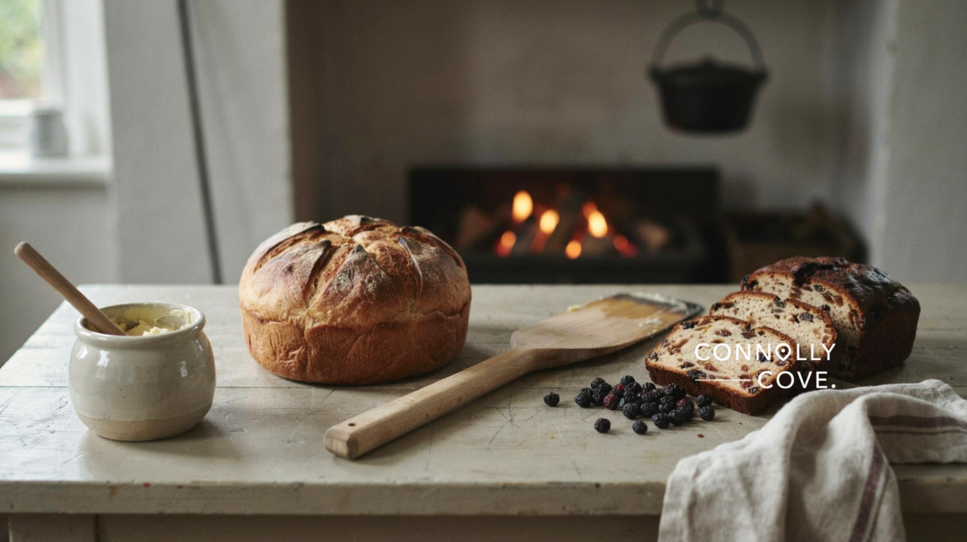 A rustic loaf of bread, a wooden spatula, butter, and berries are on a wooden table by a fireplace—evoking the hearty warmth of traditional Irish cuisine. The logo CONNOLLY COVE appears on the image.