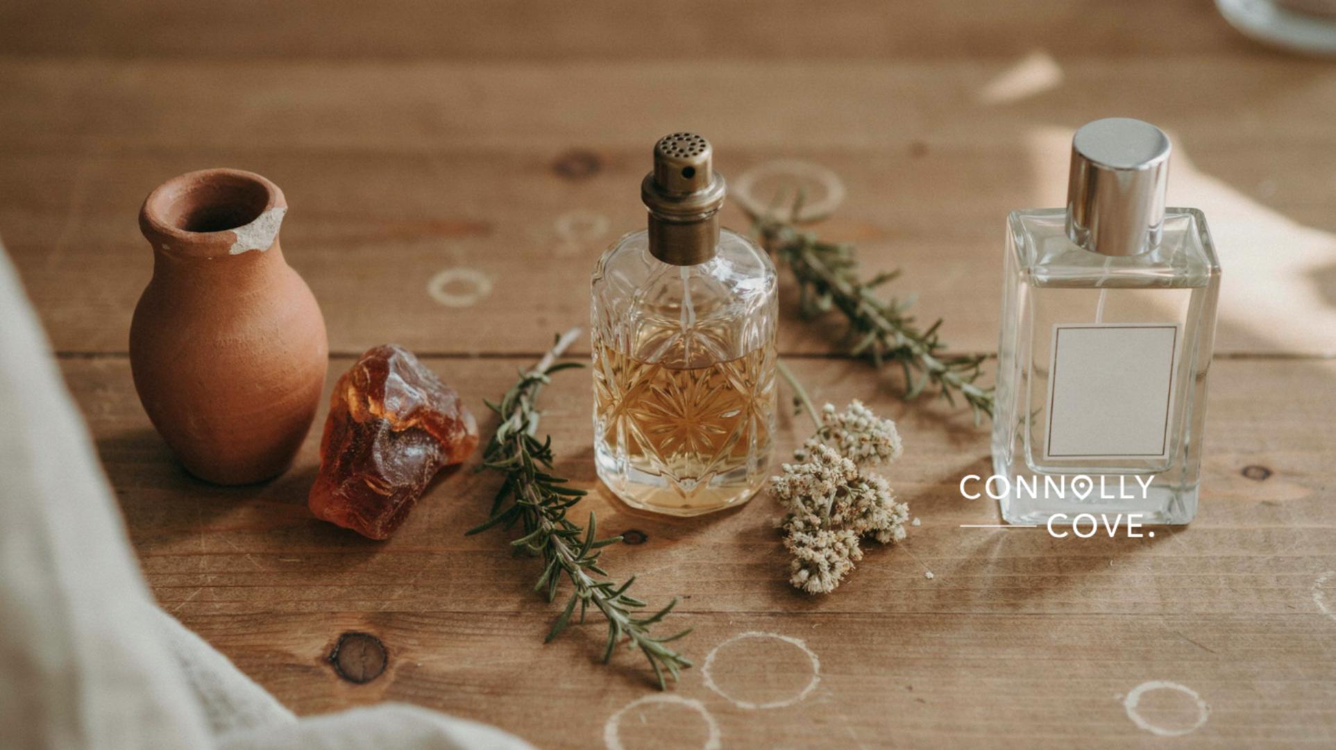 Two perfume bottles, herbs, a clay pot, and resin sit on a wooden table with Connolly Cove text. The scene has a rustic, natural feel, evoking the artistry of perfumery and the timeless allure of fragrance through the evolution of perfume.