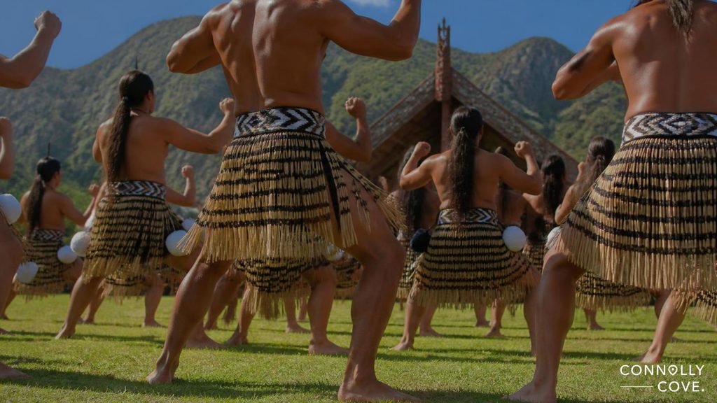 People wearing traditional Māori attire perform a cultural dance, likely a Māori Haka, on grass, with a carved wooden building and mountains in the background. The text CONNOLLY COVE. appears in the bottom right corner.