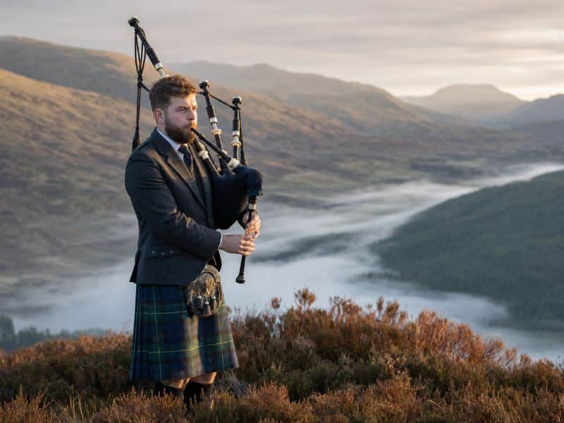 A lone piper playing the bagpipes of Scotland on a heather hillside above a misty Highland glen at dawn