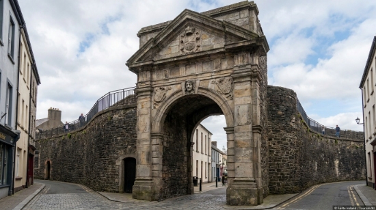 A historic stone city gate—Bishops Gate—with an arched entrance stands between two curved stone walls, marking a grand entryway into the walled city. Flanked by narrow streets and buildings under a cloudy sky, it exudes centuries of heritage and character.