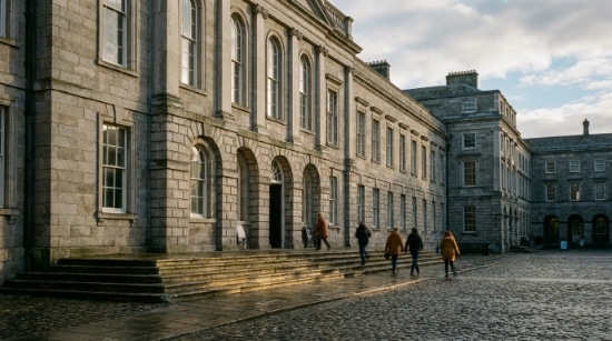 People walk up steps towards a large stone building with arched doorways and tall windows in a cobbled courtyard on a cloudy day, reminiscent of the grandeur often associated with sites that house treasures like The Book of Kells.