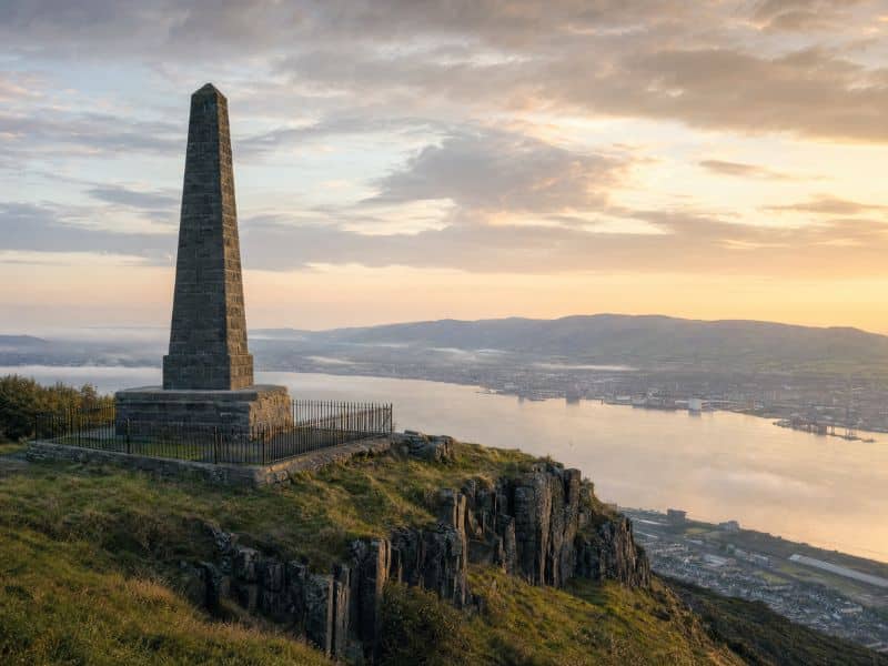Illustration evoking the Knockagh Monument at sunrise, a tall stone obelisk on a basalt cliff above Belfast Lough