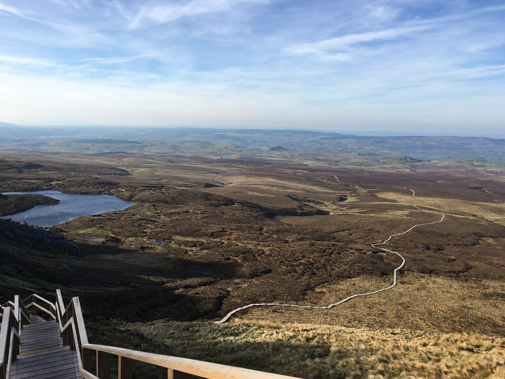 A wooden staircase leads down to a vast, open Fermanagh moorland, with a winding boardwalk path and a small lake under a blue sky with light clouds.