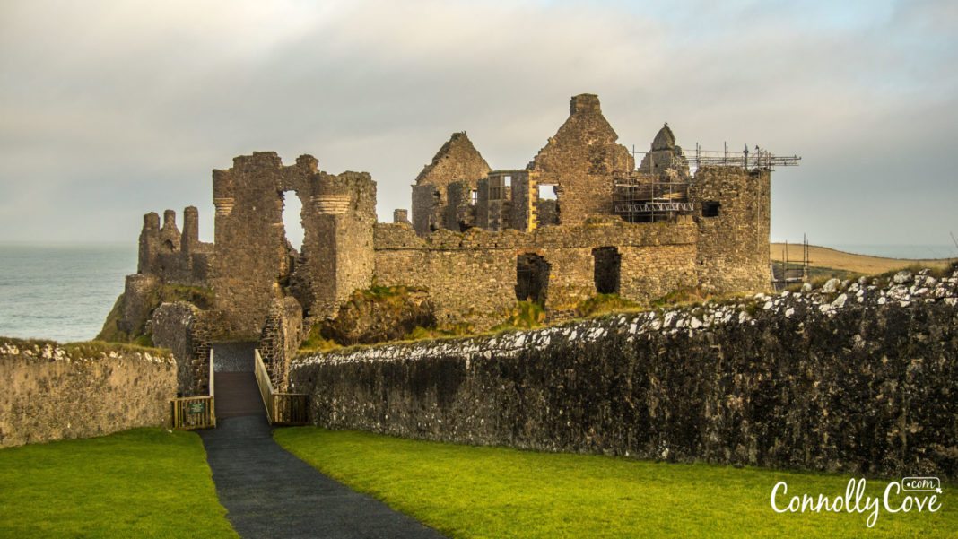 Ruins of Dunluce Castle stand atop a cliff overlooking the sea, with stone walls, scaffolding, and a path leading toward the entrance.
