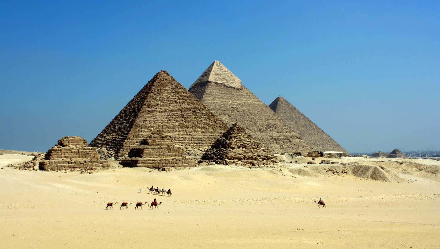 The Pyramids of Giza rise from the sandy desert under a clear blue sky, with groups of people riding camels in the foreground.