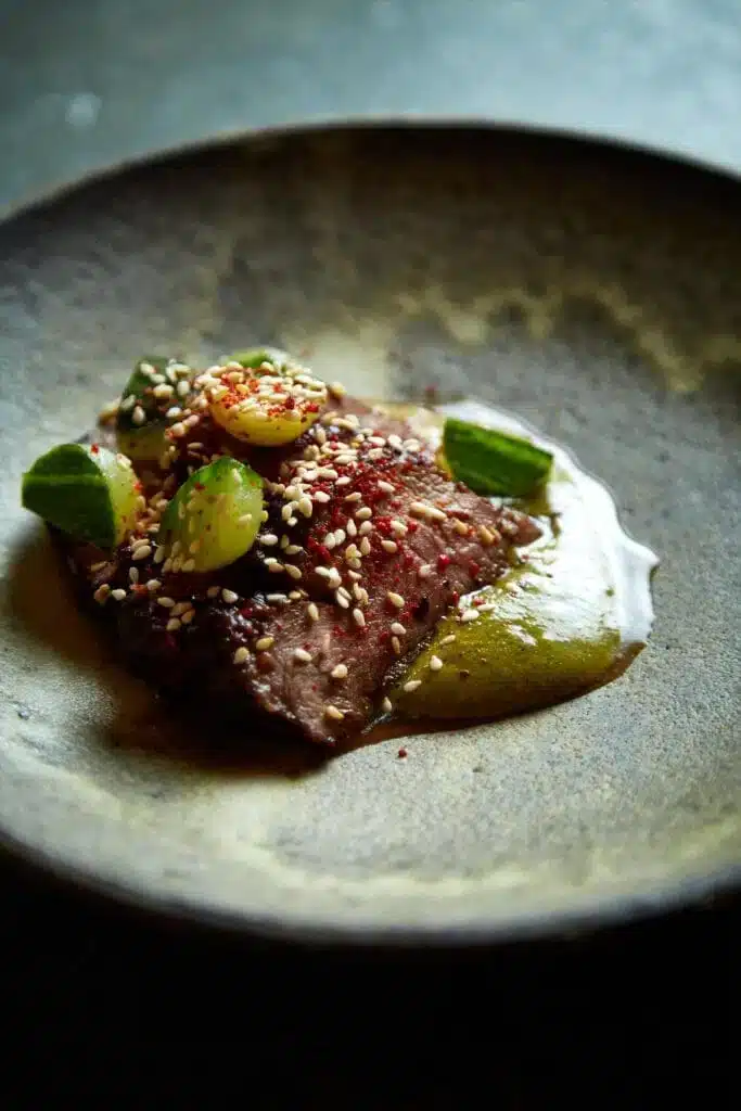 A plated dish featuring a piece of seared meat garnished with sauce, cucumber slices, sesame seeds, and fresh herbs on a ceramic plate.