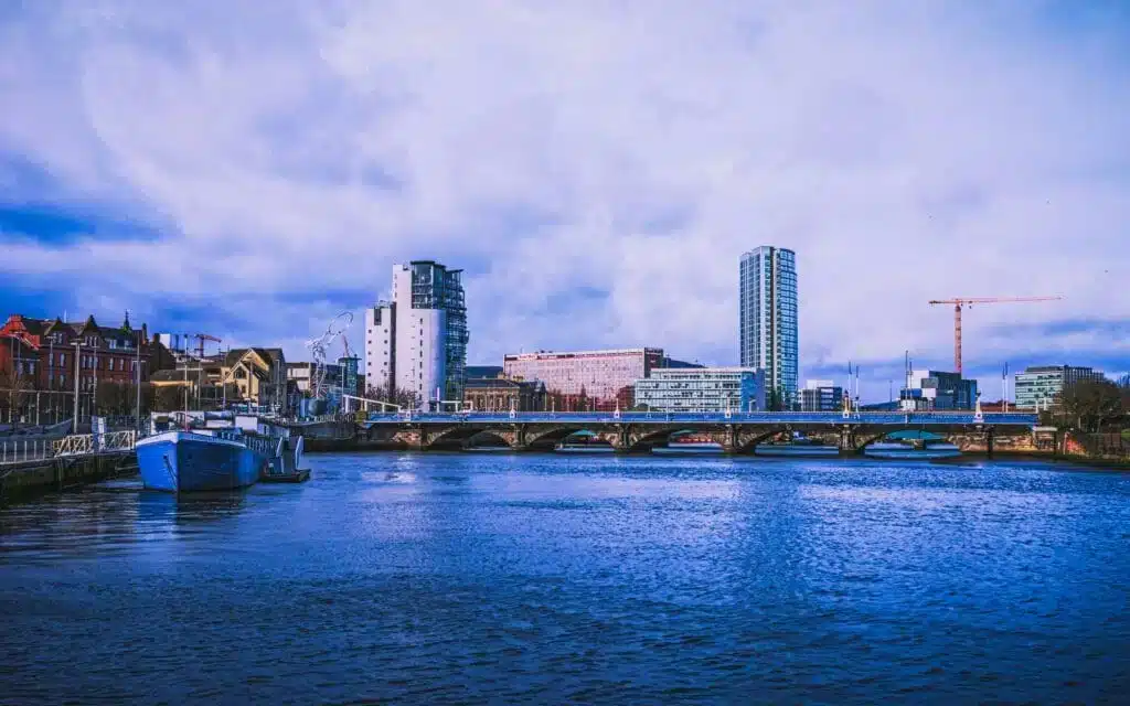 A cityscape view of Belfast featuring modern buildings, a bridge spanning the River Lagan, and a docked boat under a partly cloudy sky.