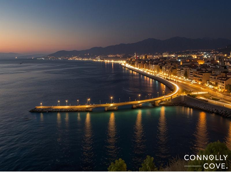 A coastal city at dusk in Italy with streetlights illuminating a curving shoreline road beside calm blue water, mountains in the background, and buildings lit up along the coast—Messina's beauty shines. Connolly Cove is written in the corner.
