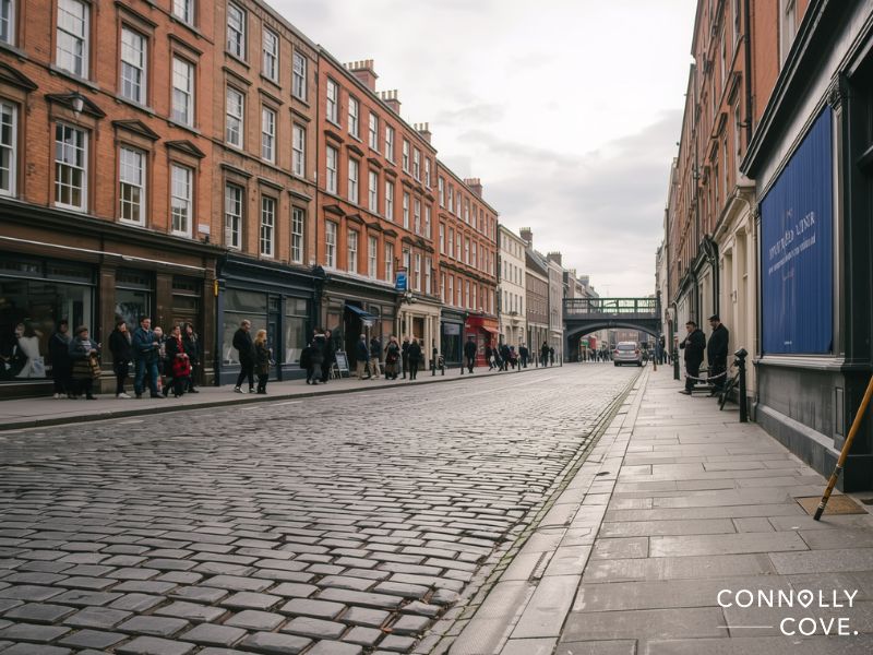 A cobblestone street lined with red-brick buildings, shops, and people walking or standing on both sides. The sky is overcast, and a bridge crosses the street in the distance—perfect for your Dublin holiday. Connolly Cove logo is in the corner.
