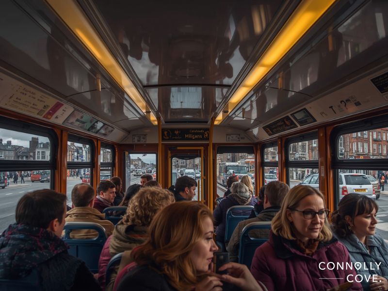 Passengers sit inside a city tram with large windows, chatting as they travel past urban streets. Outside, buildings and cars hint at city life—perhaps even near Trinity College—while the image is branded Connolly Cove.