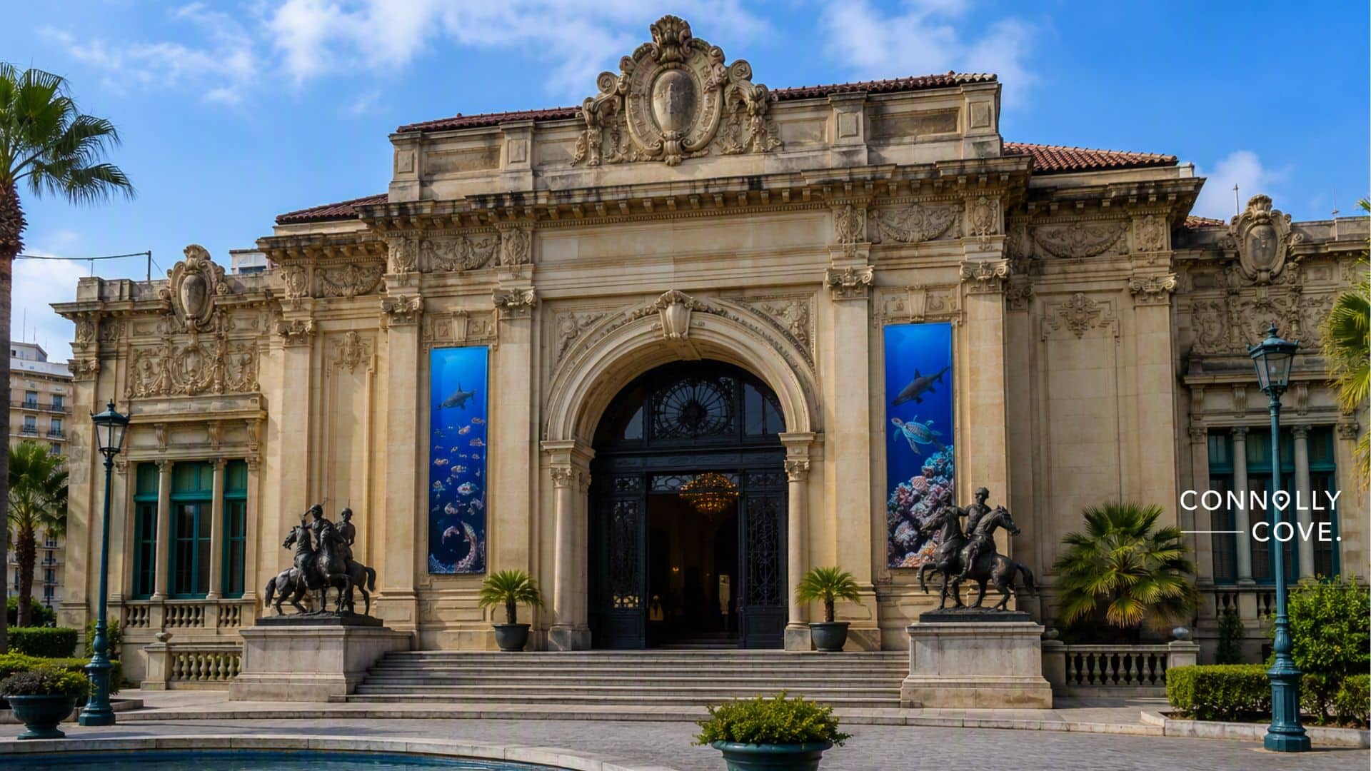 A grand historic building in Alexandria featuring ornate stonework, two statues of horse riders, a large archway entrance, palm trees, blue banners with sea life hinting at the Aquarium Museum within, and a sign reading Connolly Cove.