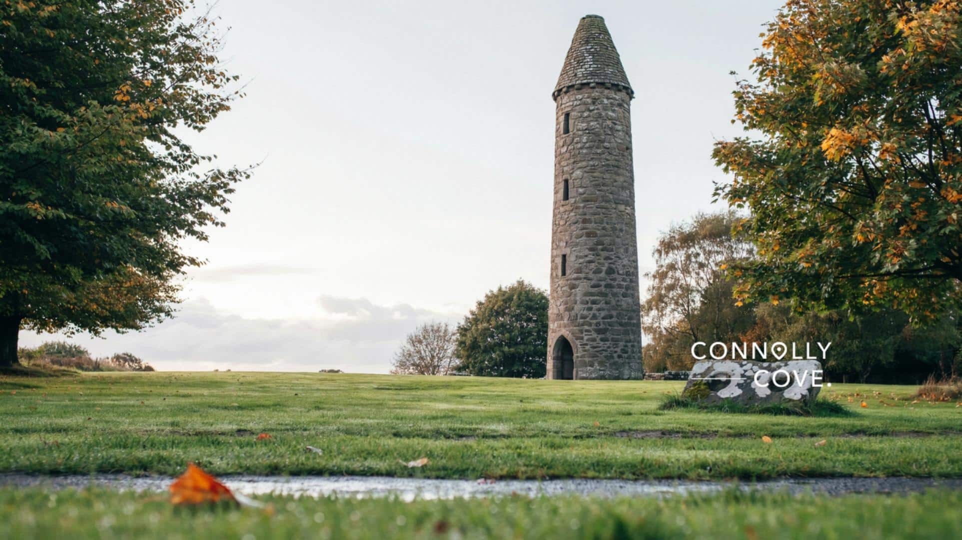 A round tower of stone stands on green grass, surrounded by autumn trees under a cloudy sky. A rock with Connolly Cove written on it sits in the foreground.